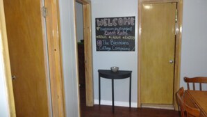 A hallway with wooden doors, a blackboard with 'WELCOME' and 'Beach Rubs' written on it, a small table with a bowl, and a wooden chair.