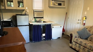 Een compacte keuken met een koelkast, een wastafel en een raam met zonwering. In de woonkamer staat een houten aanrecht en een bank met een patroon.