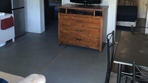 Un espace de vie compact comprenant un coin cuisine, un téléviseur à écran plat posé sur une commode en bois, une table à manger avec des chaises, et un lit à l’arrière-plan.