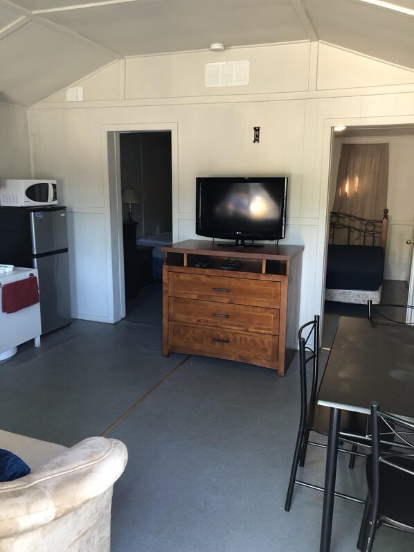 A compact living space with a kitchenette, a flat-screen TV on a wooden dresser, a dining table with chairs, and a bed in the background.