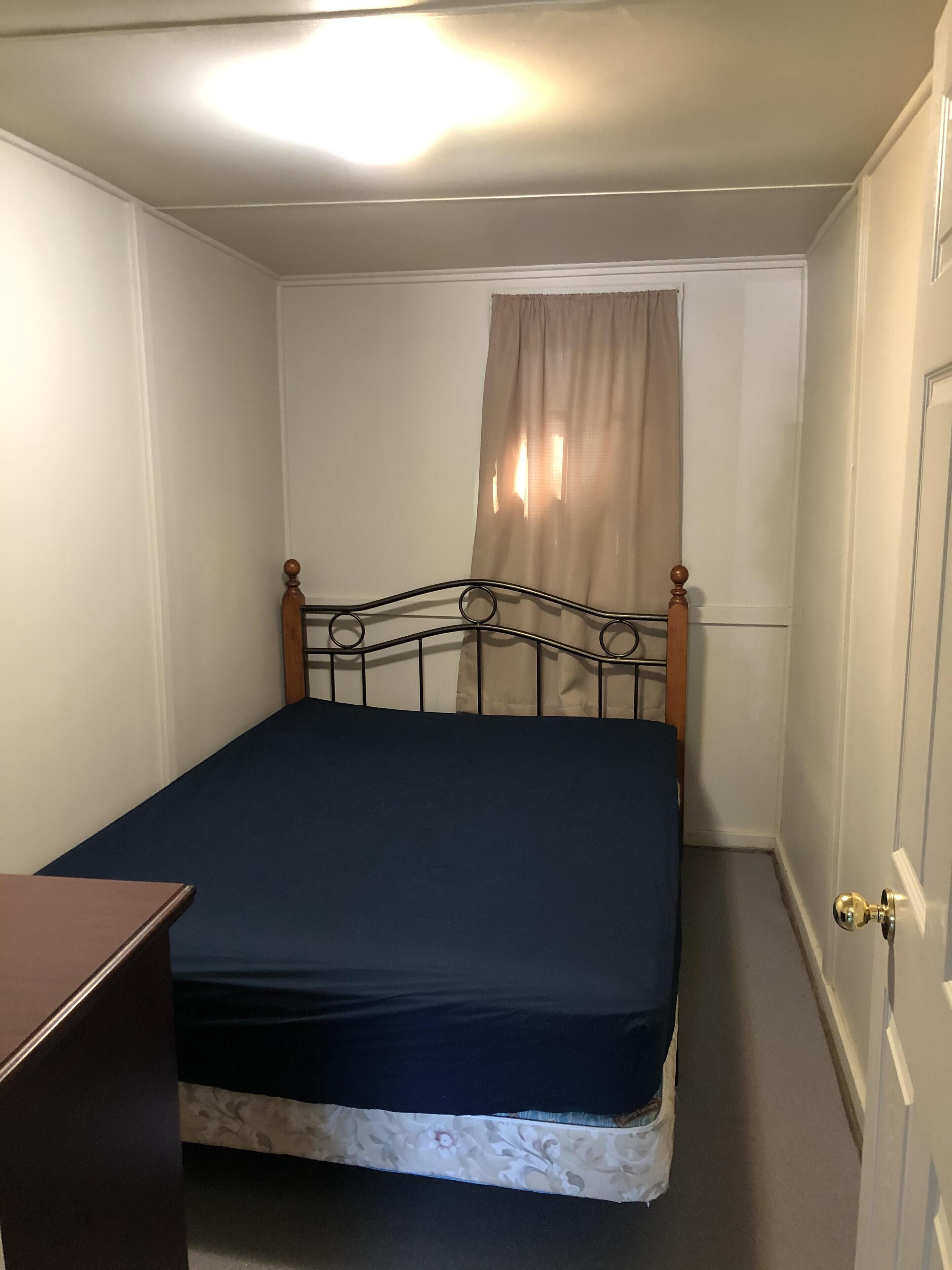 A small, single-bed room with a wooden headboard, a bedside table, and a window with curtains.