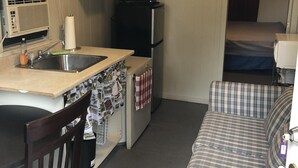 Pieni, käytännöllinen keittiönurkkaus, jossa on pesuallas, pieni jääkaappi ja mikroaaltouuni. Olohuoneessa on tuoli ja sohva.