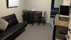 Ett kompakt rum med en svart lädersoffa, ett litet matbord med fyra stolar, en vit TV som är monterad på väggen och en liten köksdel med en diskho och en arbetsbänk.