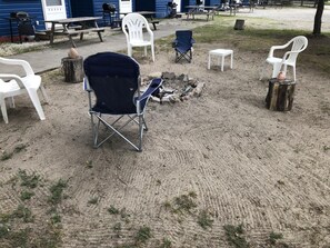 Un espace extérieur aménagé avec des sièges, comprenant un foyer, entouré de chaises en plastique blanches et d’une chaise pliante bleue.