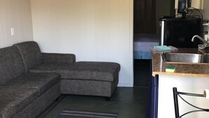 A compact living space with a sofa, a small table, a chair, a kitchen area with a sink, and a television. - Dock Life Cottage 8 - Riverside, 1 Bedroom, Wasaga (Wasaga Beach)