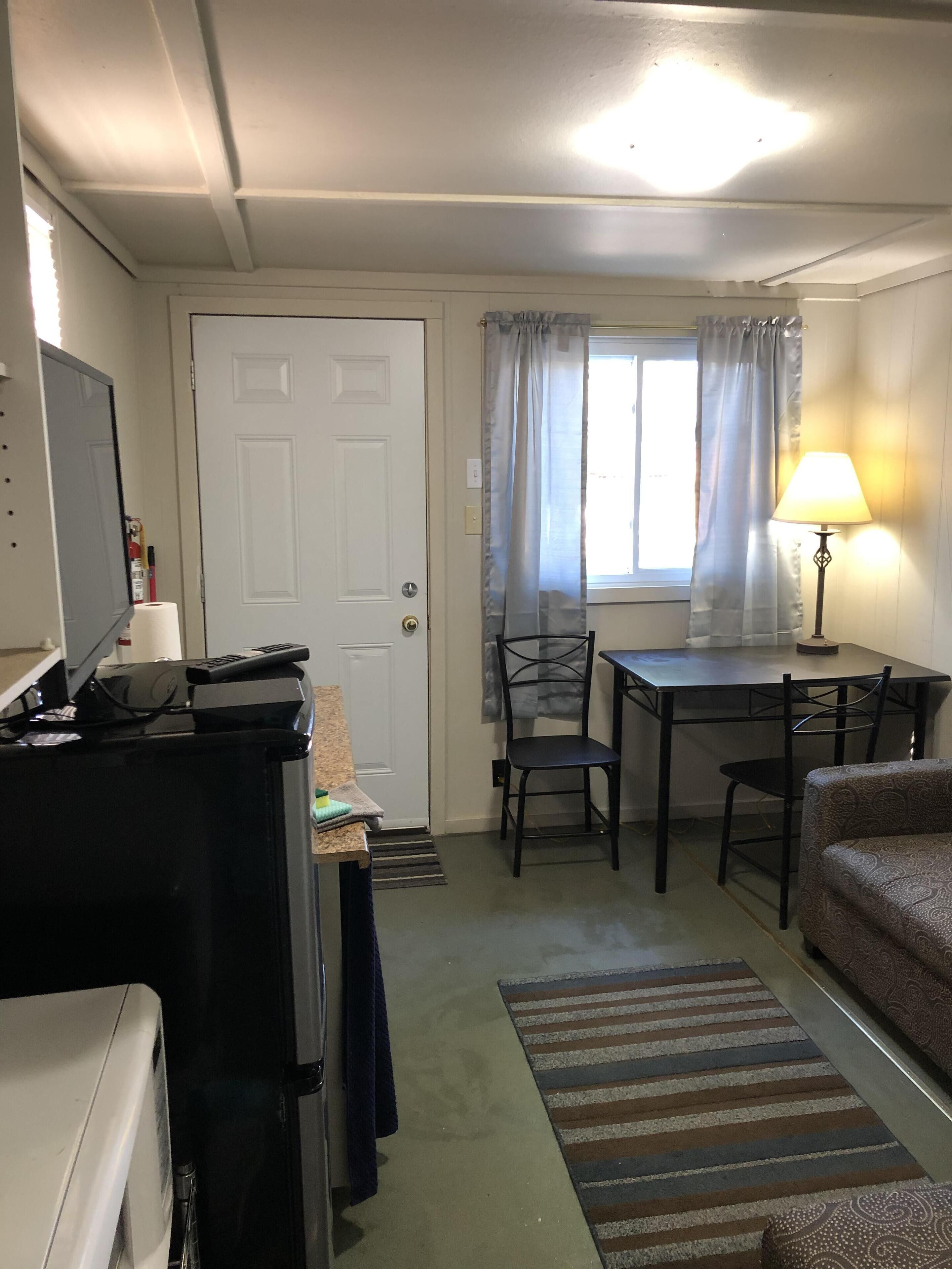 Une pièce compacte comprenant un coin cuisine, une table à manger avec des chaises, un canapé et une fenêtre avec des rideaux.
