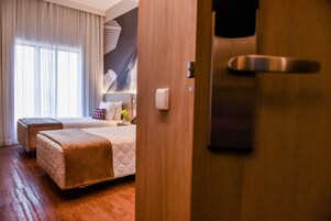 Quarto de hotel com duas camas, uma janela grande com cortinas, mesa de cabeceira com abajur e porta com puxador.