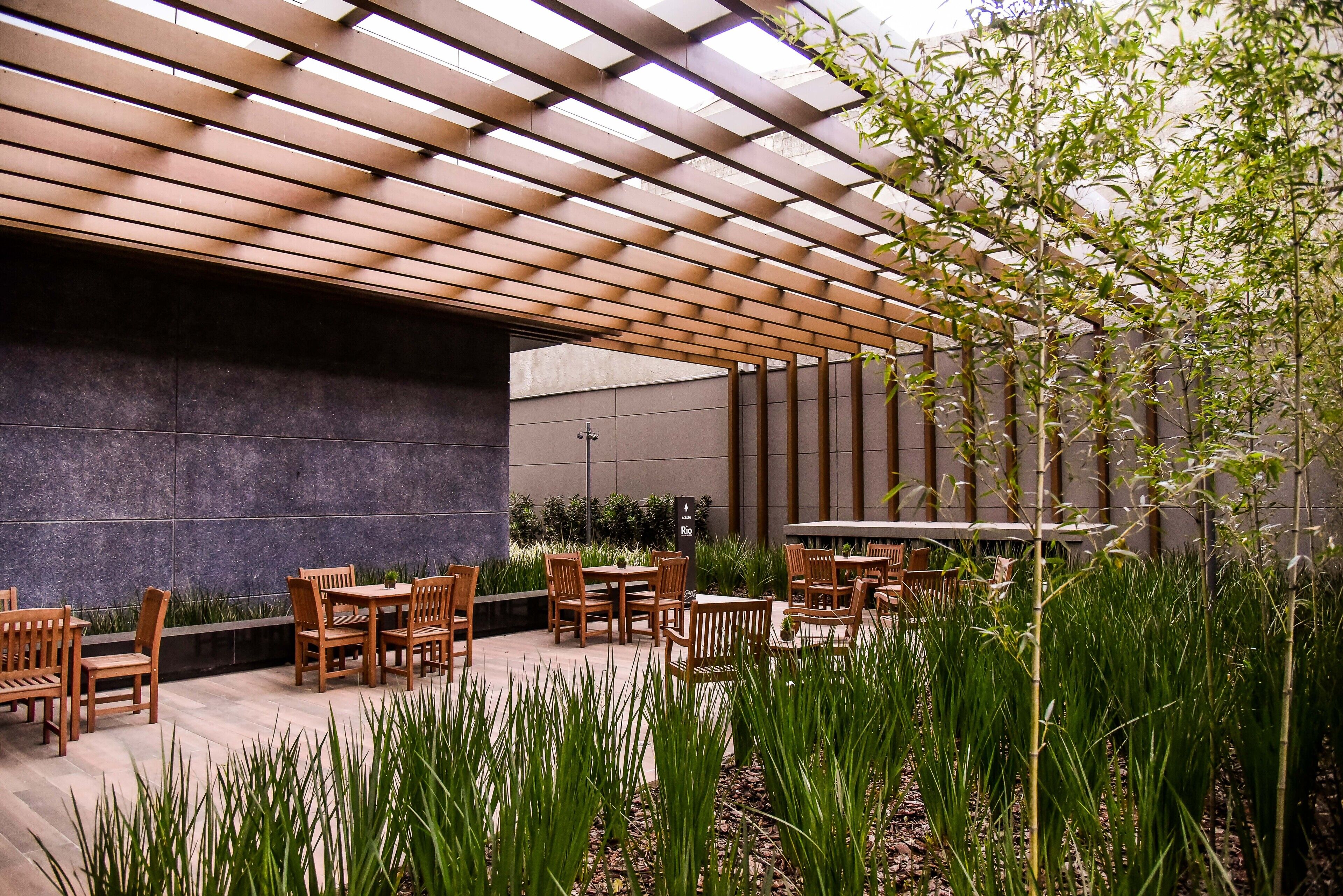 Un espacio al aire libre con mesas y sillas de madera, rodeado de vegetación y una estructura de pérgola.