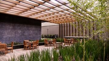 Un espacio al aire libre con mesas y sillas de madera, rodeado de vegetación y una estructura de pérgola.