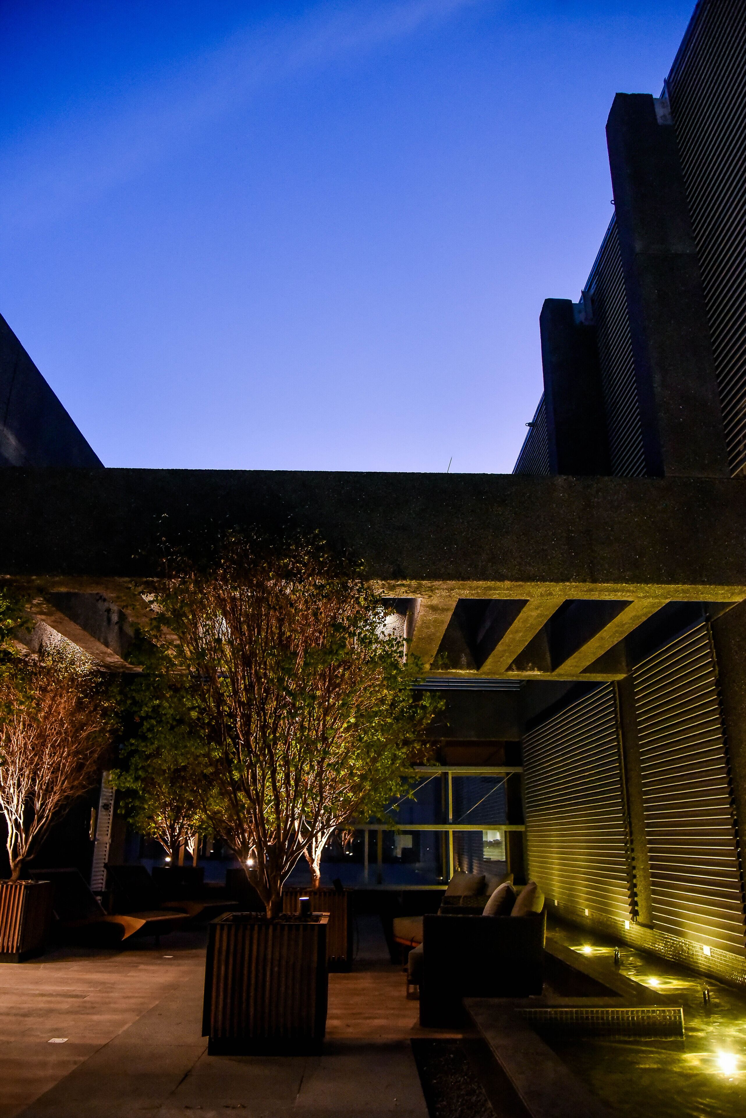 Un espace extérieur aménagé avec des jardinières et un élément décoratif aquatique, situé sous une structure couverte et offrant une vue sur la façade du bâtiment.