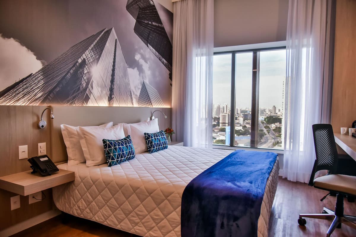 A hotel room with a large bed, a desk, a chair, a phone, and a city view.