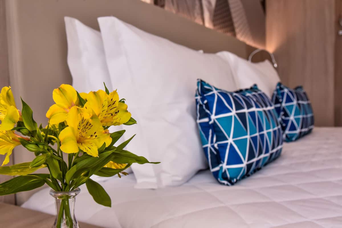 A bed with white and blue patterned pillows, a clear glass vase with yellow flowers, and a white headboard.