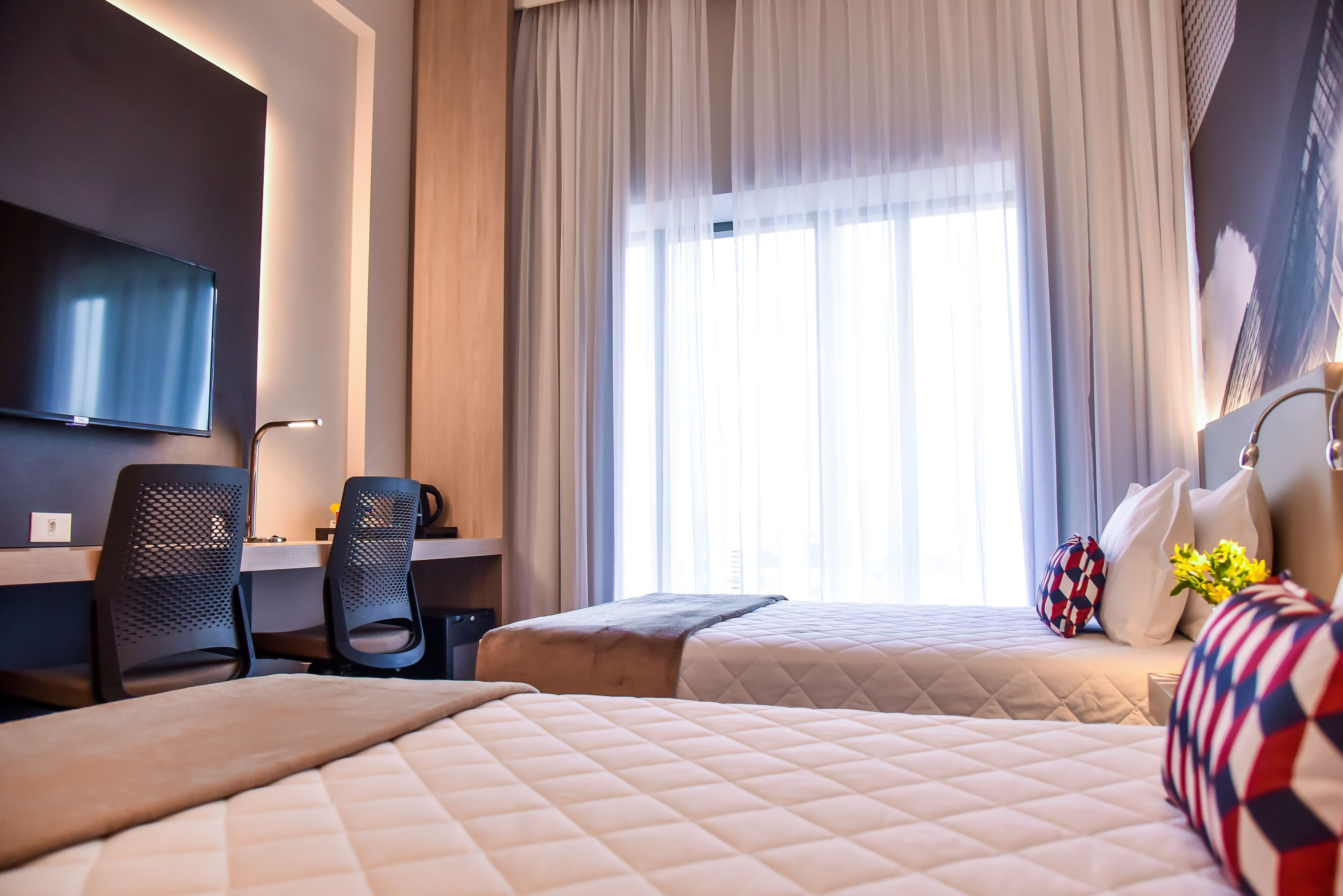 Een hotelkamer met twee bedden, een bureau, een stoel, een televisie en een groot raam met gordijnen.