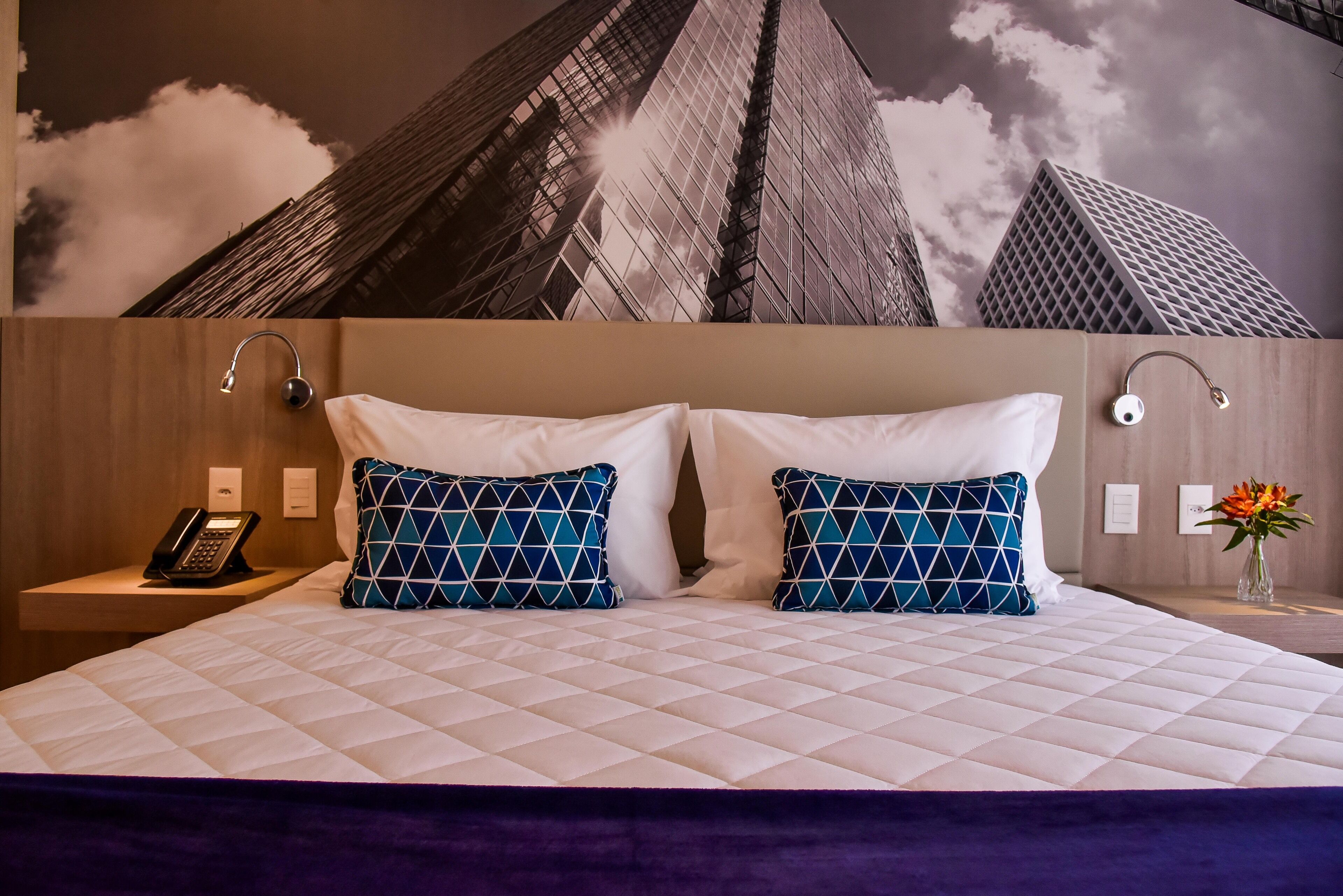 Habitación de hotel con una cama grande, dos almohadas estampadas, una mesita de noche con un teléfono y un mural en la pared con rascacielos.
