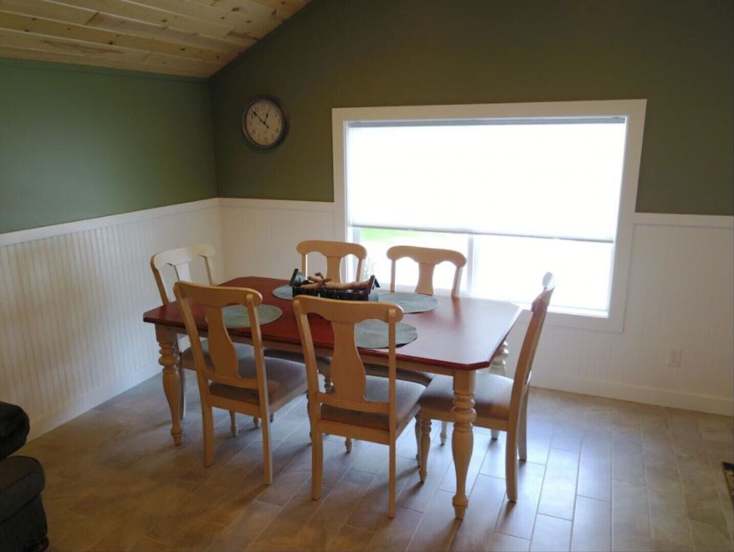 Un comedor con una mesa de madera y cuatro sillas, un reloj en la pared y una ventana que deja entrar luz natural.
