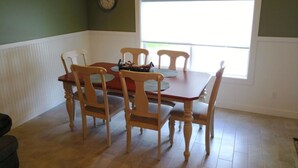 A dining area with a wooden table and four chairs, a clock on the wall, and a window providing natural light.