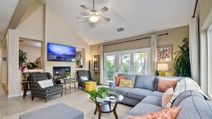A spacious living room with a grey sofa, ottoman, and a glass coffee table. A television is mounted on the wall, and there is a ceiling fan. - Two Homes, One
 Lot Sleeps 18, Pool & Hot Tub Private Villa, 9 miles from beach. (Vista)