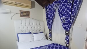 Un dormitorio con un cabecero capitonado, una cama con colcha estampada, una cortina azul y blanca y un cuadro en la pared.
