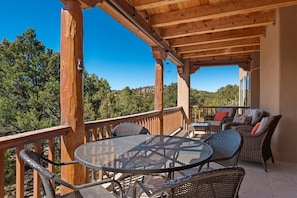 Une terrasse extérieure couverte, dotée d’une table ronde en métal et de chaises en osier, qui offre une vue sur une zone boisée.