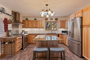 A kitchen with wooden cabinets, a stainless steel refrigerator, a central island with stools, and a hanging light fixture. - Sunlit Pines - Tucked in the Trees, Private Escape, Easy Drive to Plaza - NEW Listing (Santa Fe)