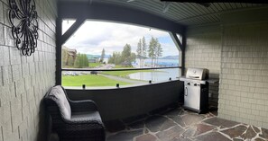 A covered outdoor area with a stone floor, a grill, a seating area with a cushioned bench, and a view of a lake and buildings. - Best spot on the Lake, Water Front, View of the floating Green Golf Coarse (Harrison)