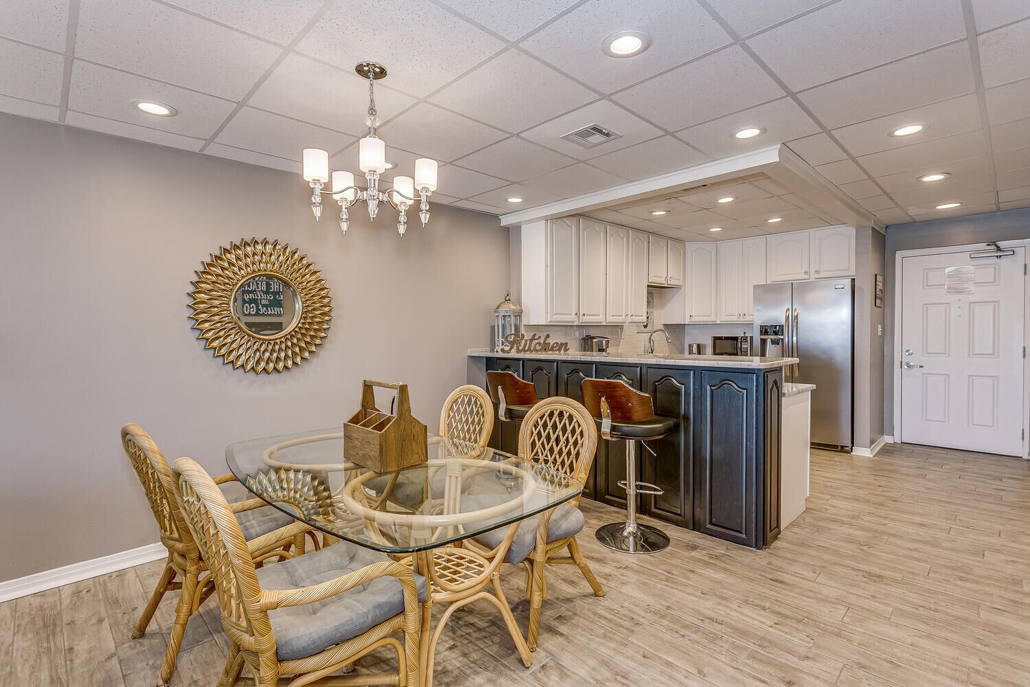 A modern kitchen with white cabinets, a stainless steel refrigerator, and a glass dining table with wicker chairs.