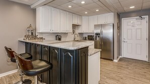 A kitchen with white cabinets, a stainless steel refrigerator, a marble countertop, and two bar stools. - Directly on the Ocean! Beautiful Views! Free Beach Chairs and Umbrella sleeps 6 (Pensacola Beach)