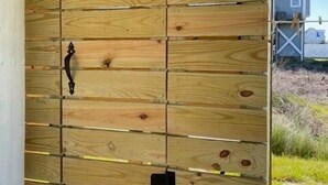 Eine Wand aus Holzpaneelen mit einem schwarzen Griff, vermutlich eine Schiebetür, in einem überdachten Außenbereich mit einer Bank und einem Haus im Hintergrund.