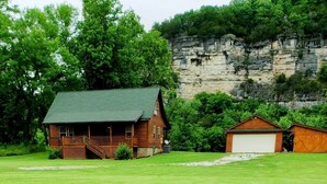 A rustic cabin with a green roof, a porch, and a garage. It is surrounded by lush greenery and a rocky cliff.