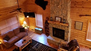 A cozy cabin interior with a stone fireplace, wooden walls, a leather sofa, and a television.