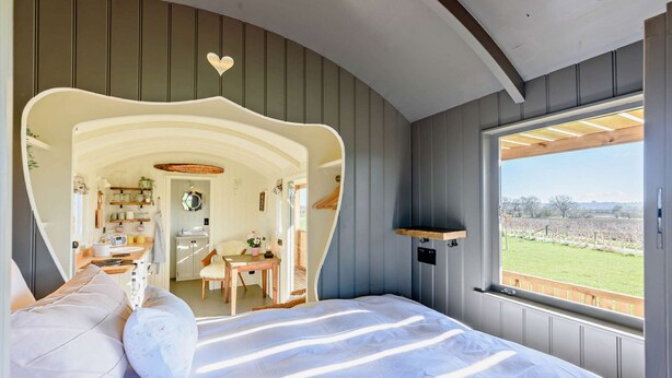 A bedroom with a curved headboard, a large window with a view of a vineyard, and a small wooden table with a lamp. - Luxury shepherd's hut for 2 in the Cotswolds - Aurora (Faringdon)