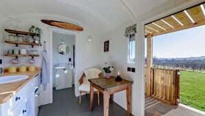 A cozy kitchen with wooden shelves, a white sink, and a dining area with a table and chairs. - Luxury shepherd's hut for 2 in the Cotswolds - Aurora (Faringdon)