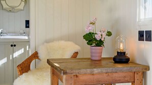 A cozy room with a wooden table, a wicker chair, a fluffy white chair, a pink flower pot, and a vintage lamp. - Luxury shepherd's hut for 2 in the Cotswolds - Aurora (Faringdon)
