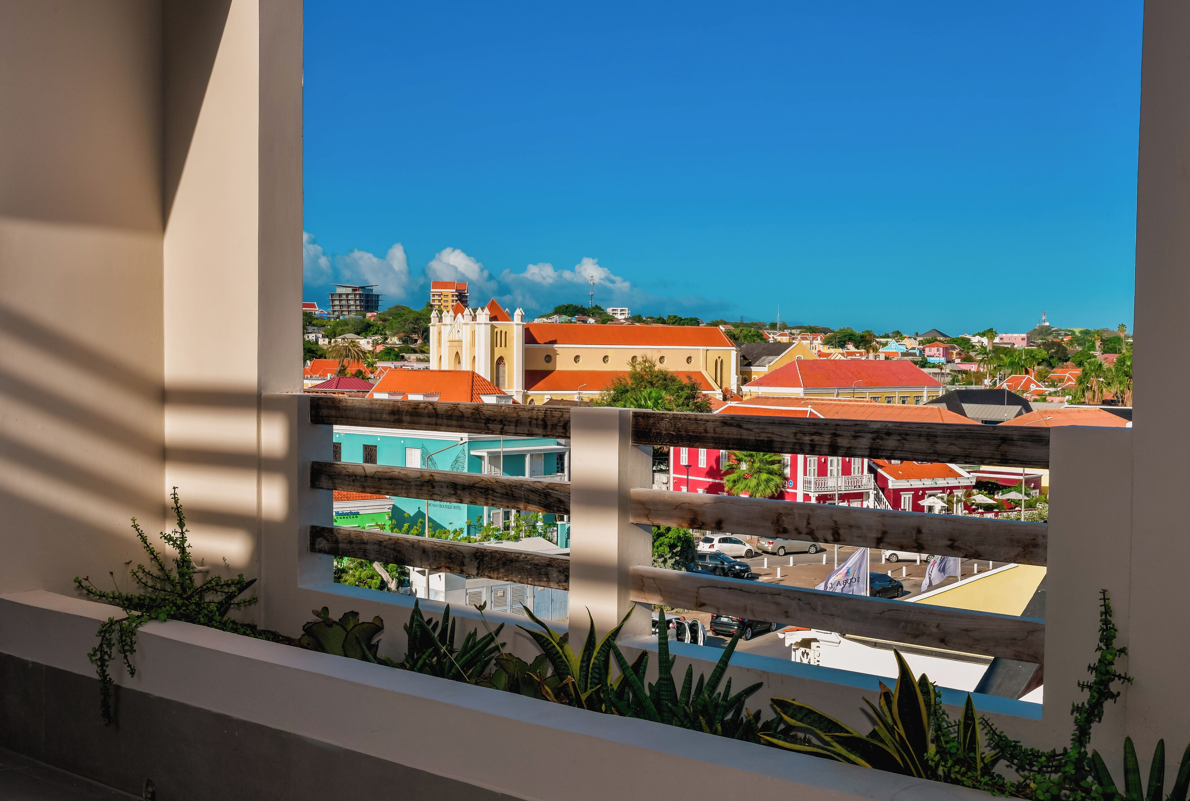 A balcony with a view of a town, including buildings with red roofs and a church with a bell tower.