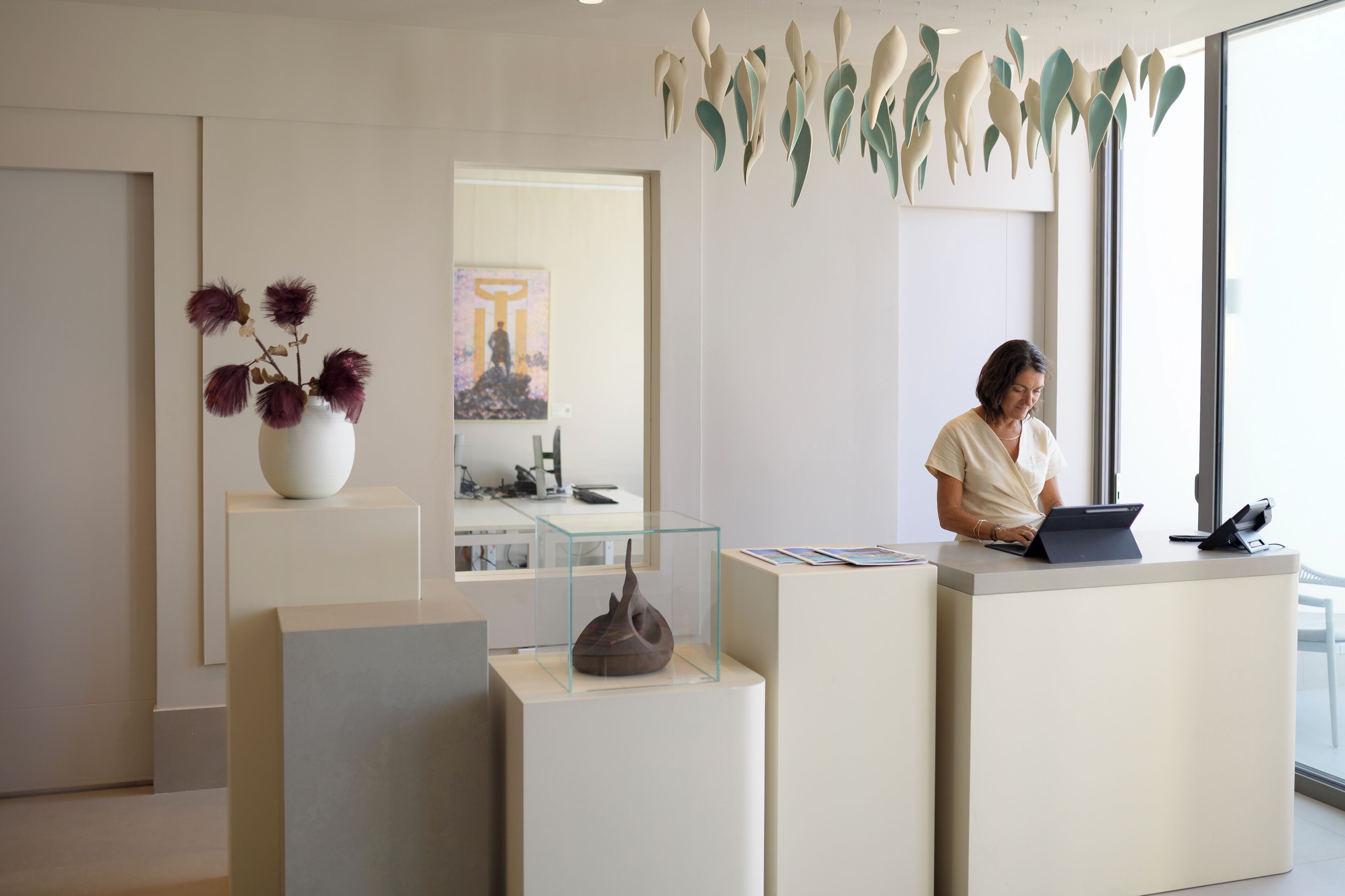 A modern reception desk with a receptionist, a vase with dried flowers, and a sculpture on display.
