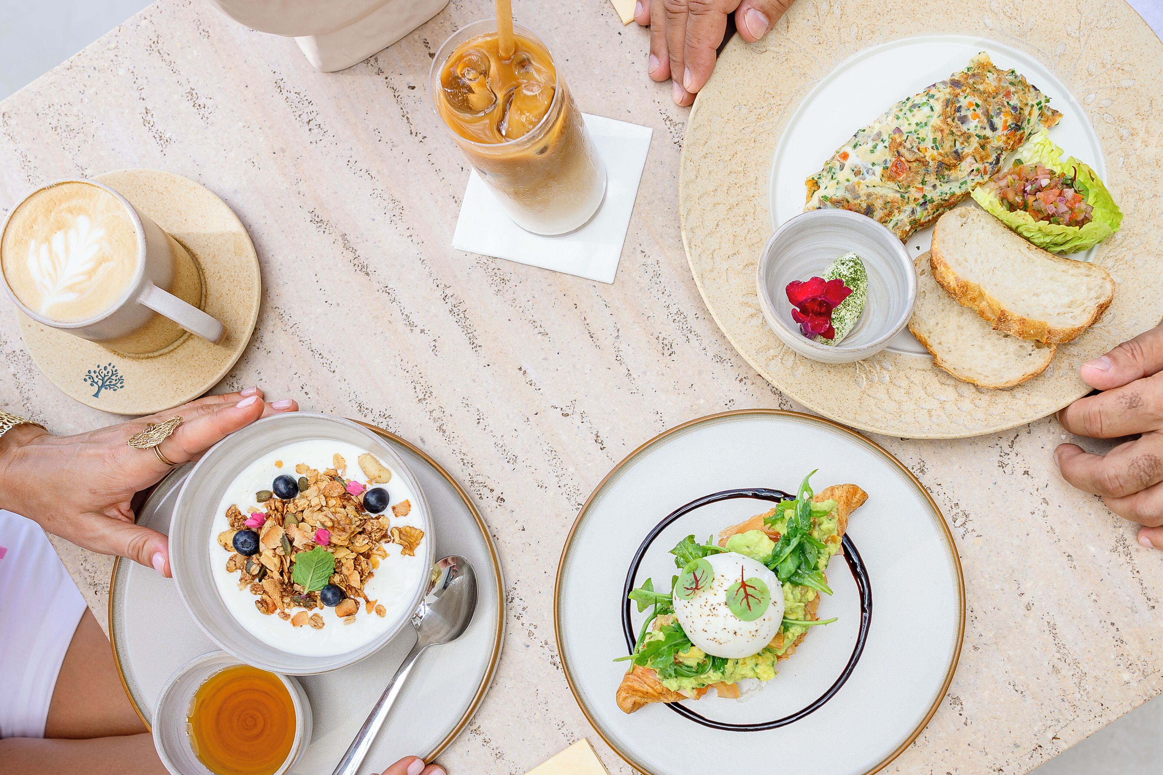 Una mesa con diversos platos, incluyendo un latte, un vaso de café helado, una ensalada y un plato con pan y un recipiente pequeño con salsa.