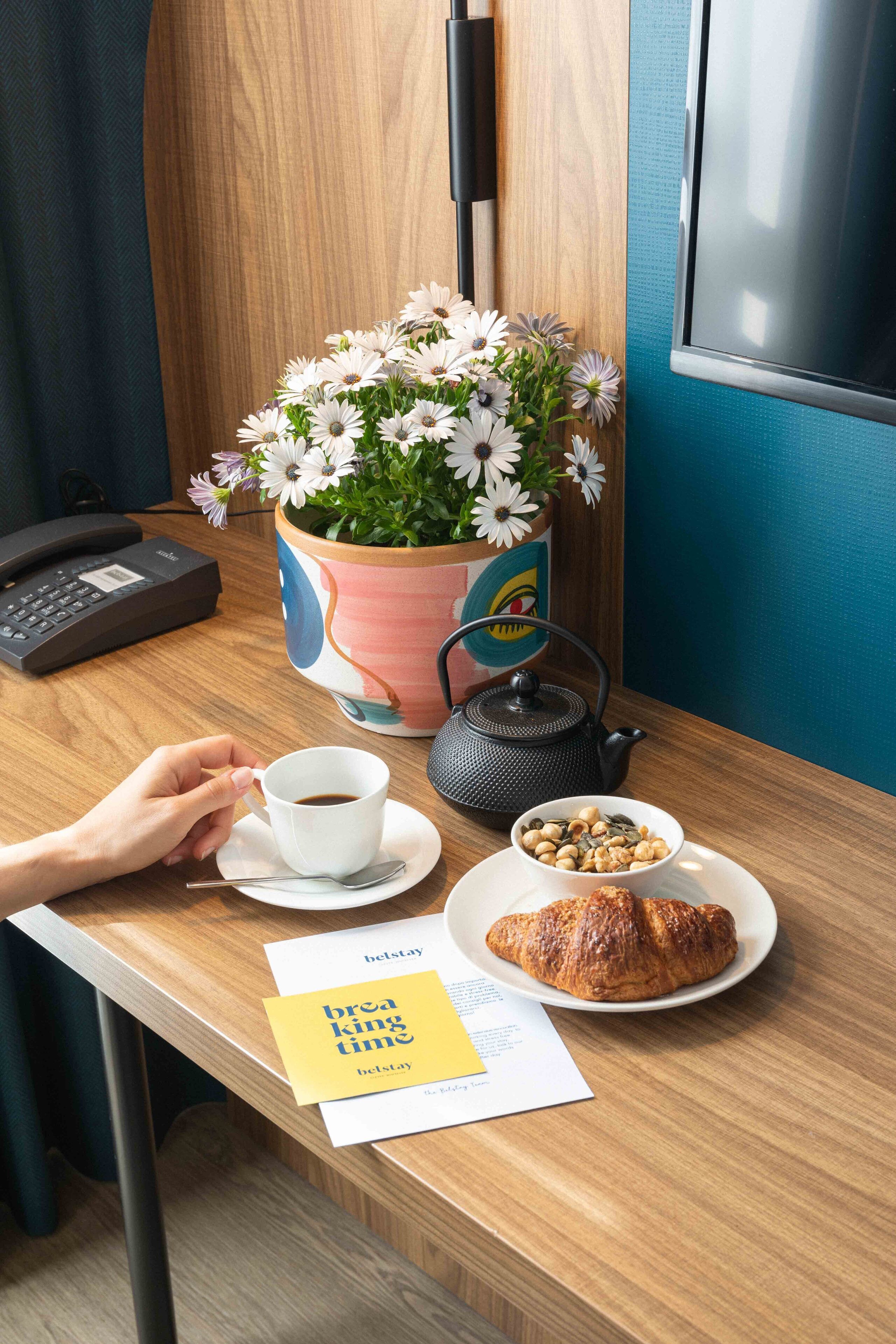 Escritorio de madera con una taza de café, un croissant, un cuenco de nueces, una tetera y una planta en maceta con flores.