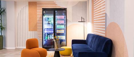 A modern lounge area with a blue sofa, orange armchairs, and a yellow ottoman. A black coffee table with a vase of flowers is in the center. Behind the sofa, there is a refrigerator stocked with beverages. The walls have a striped pattern, and the floor has a geometric tile design.