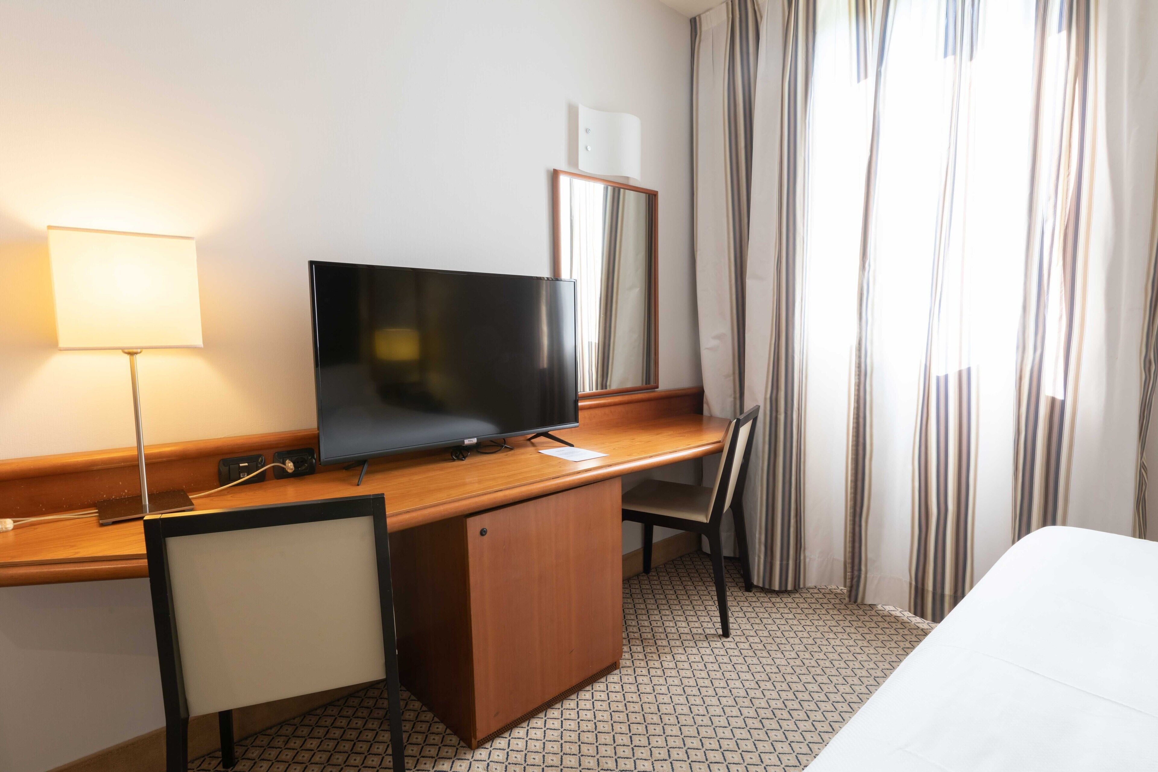 A hotel room with a wooden desk, a television, a chair, a bed, and a window with curtains.