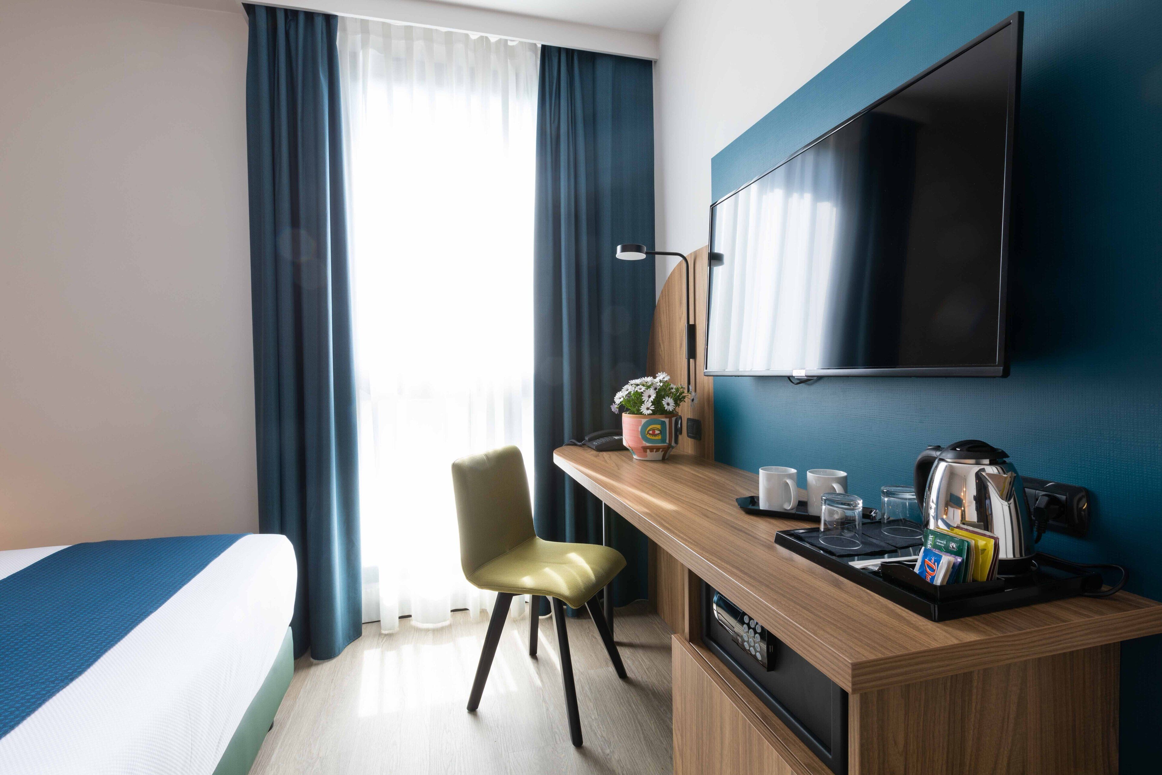 A hotel room with a bed, a desk with a coffee maker, a chair, and a TV mounted on the wall.