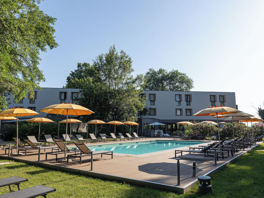 Un área de piscina con sillones de descanso y sombrillas, una piscina y un edificio al fondo.