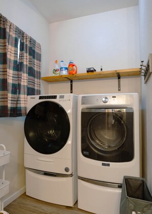 A laundry room with two front-loading washing machines, a shelf with cleaning supplies, and a plaid curtain. - East Lodge – Spacious 3BR Stay in Mobridge (Mobridge)