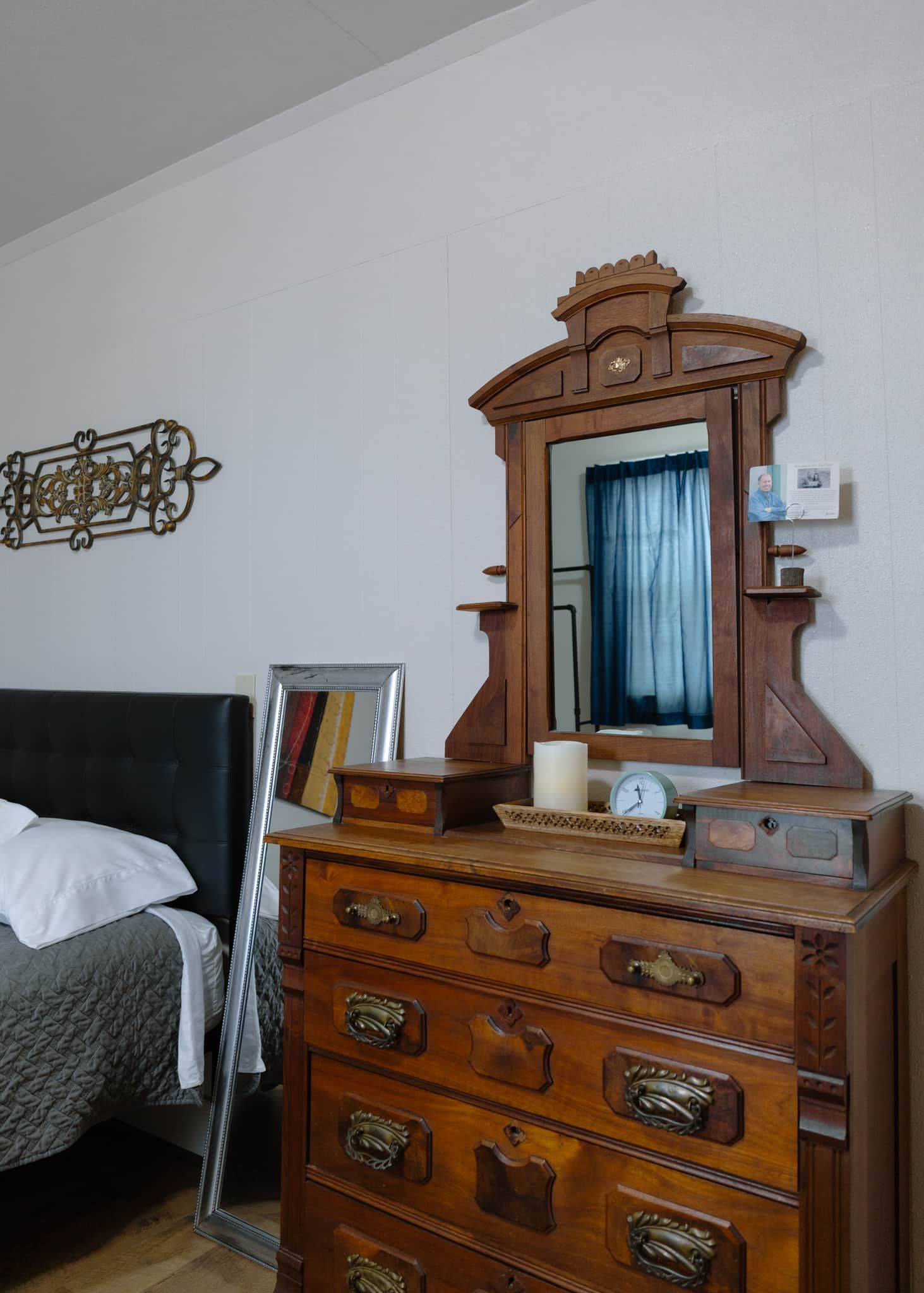A wooden dresser with drawers and a mirror, a bed with a headboard, and a wall-mounted decorative piece.