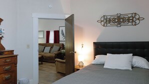 Un dormitorio con cama, mesita de noche, cómoda y puerta que da a un salón con sofá y cortinas.