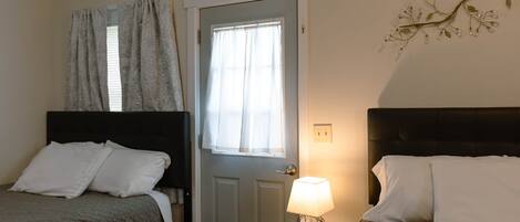 A hotel room with two beds, a door with a window, a small table with a lamp, and a wall decoration.