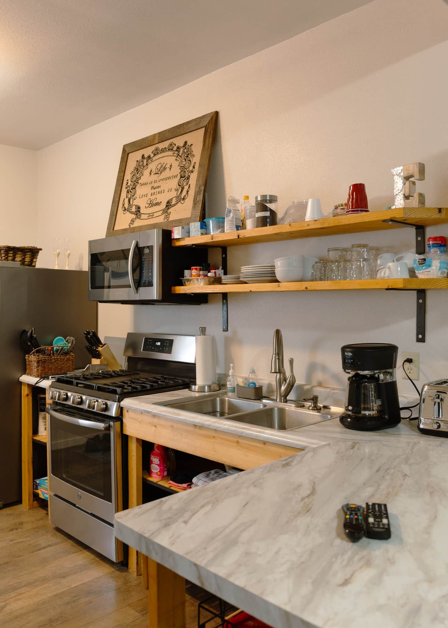 Une cuisine équipée d’un four encastré, d’un four à micro-ondes et d’une machine à café. Des étagères en bois, situées au-dessus de l’évier, servent à ranger divers ustensiles de cuisine.