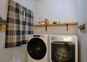 Een wasruimte met twee wasmachines, een plank met schoonmaakmiddelen en een ruitjesgordijn.