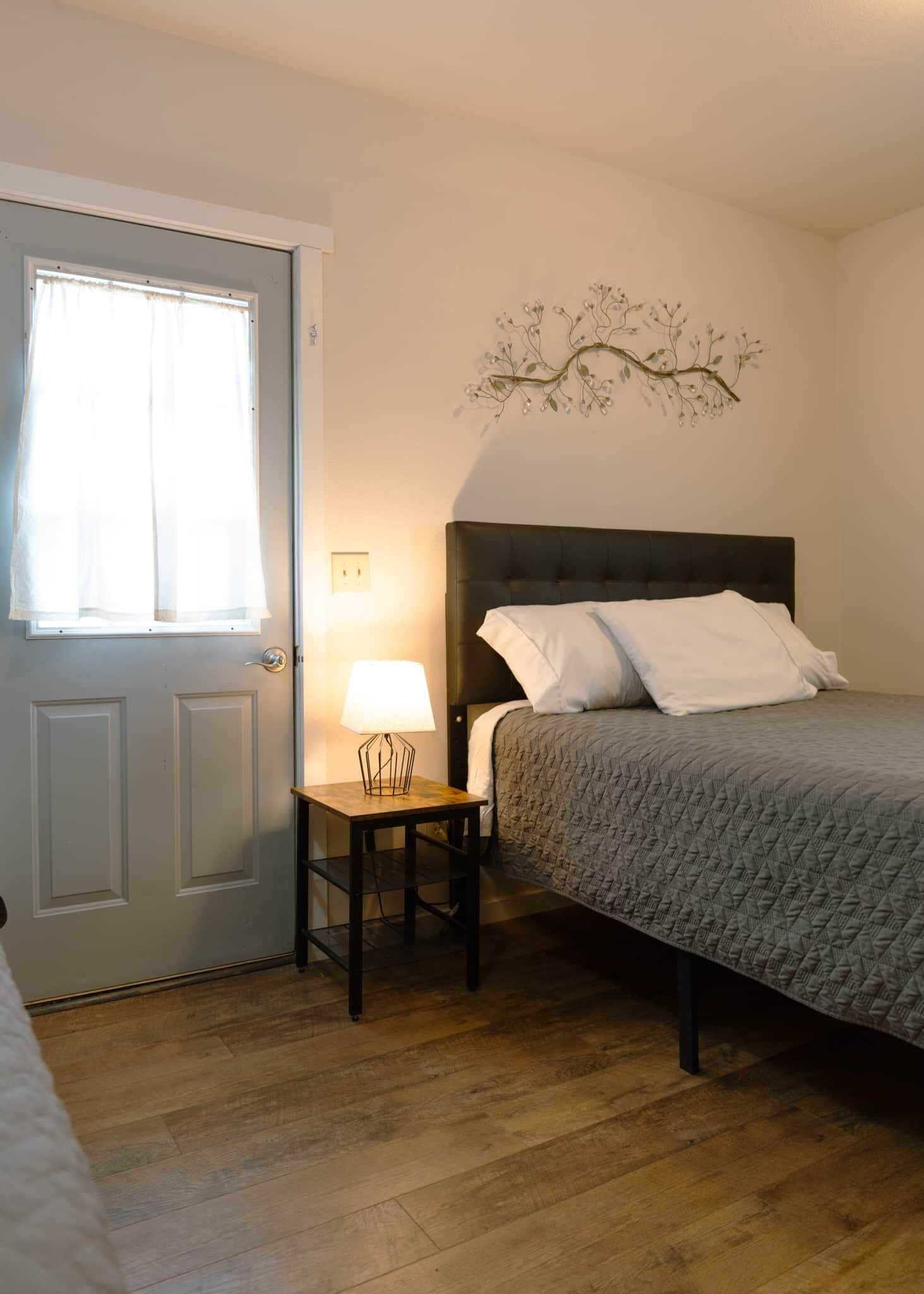 Un dormitorio con cama, una mesita de noche con l ámpara, una ventana con cortinas y un adorno en la pared.