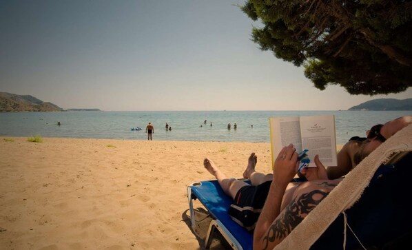 A beach scene with a person lying on a sunbed reading a book, another person sunbathing, and several people swimming in the water. - Tortuga  Pelouzo island view I (Vasilikos)