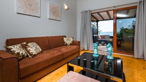 Un salon avec un canapé marron, deux coussins décoratifs, une table basse en verre et une vue sur la mer grâce à de grandes fenêtres.